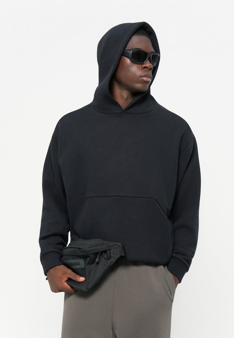 Felpa nera con cappuccio e tasca frontale, indossata con pantaloni grigi. La persona tiene una pochette nera. Occhiali da sole trasparenti completano il look.