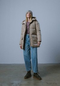 Beżowa kurtka pikowana z kapturem, szara wełniana czapka, fioletowy top i niebieskie spodnie z wysokim stanem; brązowe buty w stylu trekkingowym.