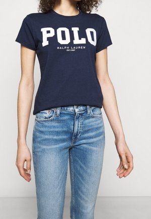 Kvinde iført en mørkeblå T-shirt med teksten "POLO Ralph Lauren EST.1967" og lyseblå højtaljede jeans mod en ensfarvet baggrund.