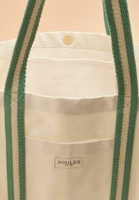 Borsa a tracolla in tela color crema con una cinghia intrecciata verde. Presenta una tasca singola e un bottone di metallo con l'etichetta "JOULES EST. 1999."
