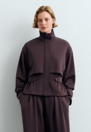 Junge Person mit kurzen blonden Haaren, die eine dunkelbraune Jacke mit hohem Kragen und geknöpften Bundlaschen sowie passende weite Hosen trägt.