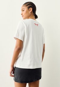 T-shirt bianca in cotone con maniche corte, con la scritta rossa "Pop Off" sul retro, abbinata a una minigonna in denim nera.