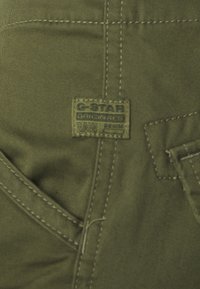 Närbild av olivgrön denimtyg med detaljerad sömnad och en G-Star Originals Raw Denim etikett.