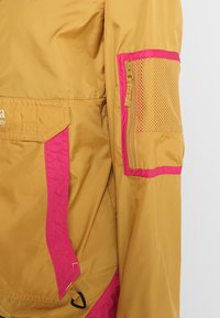 Giacca leggera gialla e rosa con tasca in rete sulla manica e tasca laterale con zip, realizzata in un materiale liscio e resistente all'acqua.