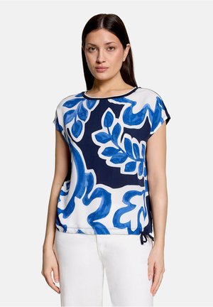 Femme portant un chemisier blanc à manches courtes avec un grand imprimé floral abstrait bleu, associé à un pantalon blanc, debout devant un fond uni.