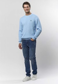 Lyseblå kabelstrikket genser med ribbede mansjetter, kombinert med mørkeblå jeans og hvite joggesko. Avslappet passform, strukturert stoff og brodert logo.