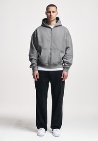 Joven de pie, vistiendo una sudadera con capucha gris con cremallera, camisa blanca, pantalones negros y zapatillas blancas sobre un fondo claro y liso.