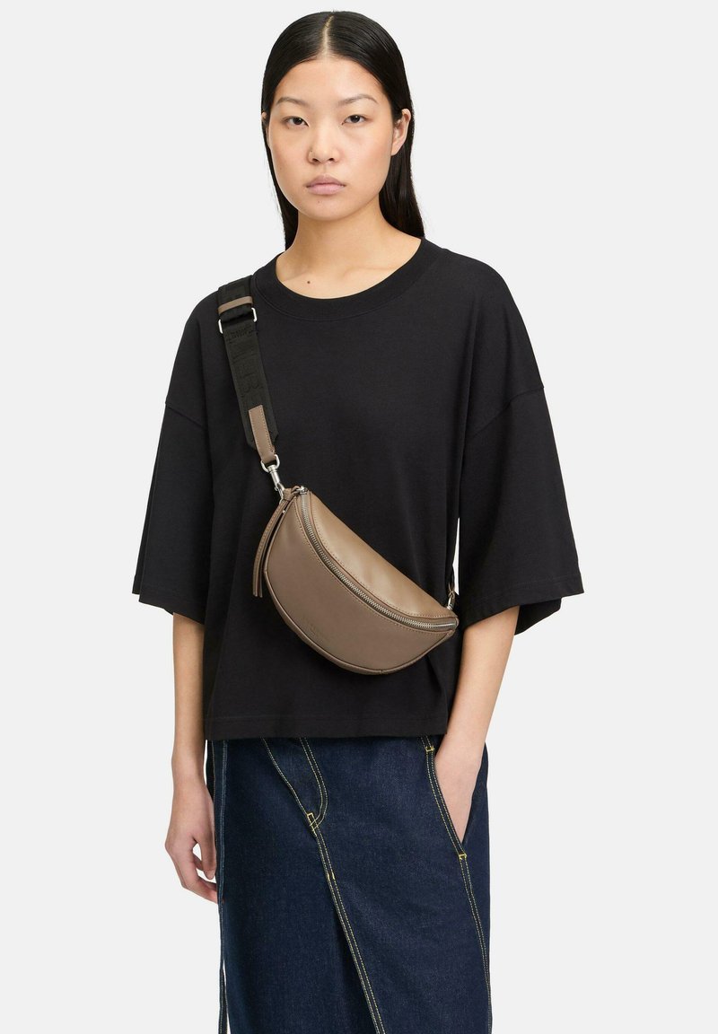 Brązowa skórzana torba na ramię o zaokrąglonym kształcie, z zamkiem błyskawicznym i regulowanym paskiem, noszona na czarnej, oversize'owej koszuli.
