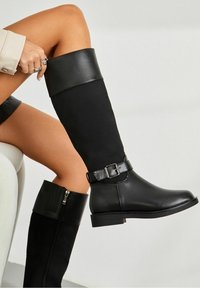Femme portant des bottes noires montantes jusqu'aux genoux, avec des garnitures en cuir et des détails de boucle, ajustant une botte en étant assise sur une chaise blanche.