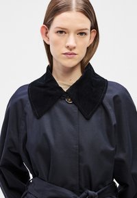 Σκούρο μπλε παλτό τύπου trench με γιακά από βελούδο κορδόνι σε μαύρο χρώμα, διαθέτει κουμπιά κλεισίματος και ζώνη στη μέση. Ομαλή υφή και δομημένος σχεδιασμός.