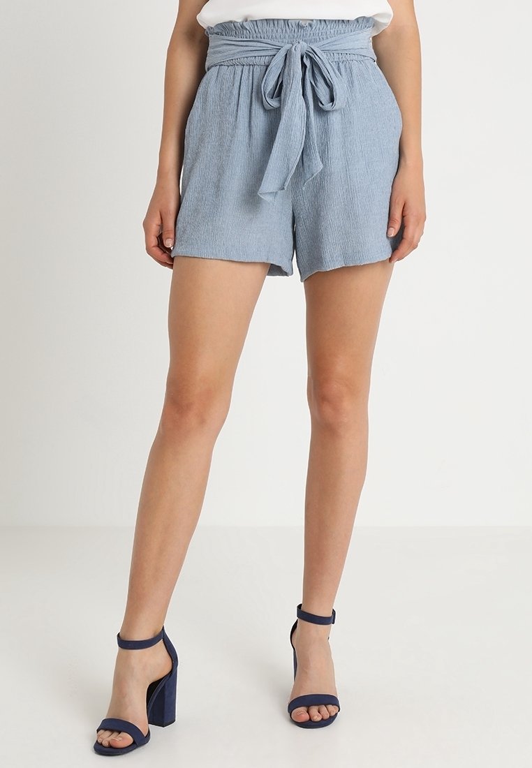 Shorts bleu clair en tissu texturé, taille élastique et ceinture nouée en tissu. Associés à des sandales à talons carrés bleu foncé avec bout ouvert.