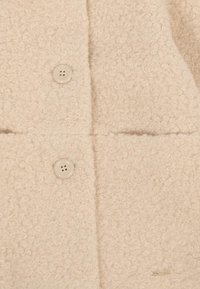 Cappotto beige con texture a tessuto ad anello, dotato di quattro bottoni, due tasche frontali e un piccolo accento metallico sull'orlo.