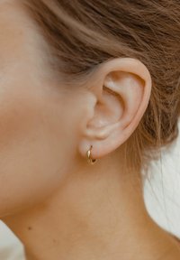 Pendiente de aro dorado en un tono de piel clara, diseño pequeño y redondo, superficie lisa, sujeto a un lóbulo, con el cabello cubriendo parcialmente la oreja.