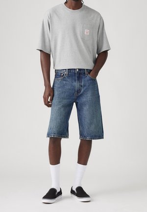 T-shirt gris à manches courtes avec une poche sur le devant, associé à un short en jean bleu. Des chaussures noires à enfiler et des chaussettes blanches arrivant à la cheville complètent le look.