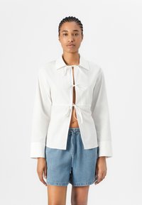 White button-up shirt with long sleeves, knot closure at the waist, and a split front. Worn with light denim shorts. Smooth cotton texture.