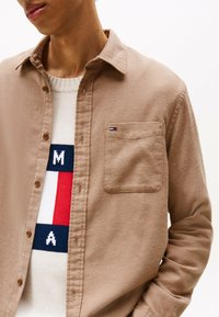 Tan, button-up shirt in a textured fabric, featuring a chest pocket and a logo patch; layered over a multicolored knitted sweater.