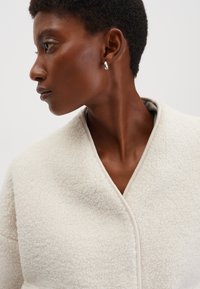 Giacca in pile bianca con texture, dall'aspetto caldo e soffice, caratterizzata da un design avvolgente unico e da un bordo a contrasto lungo i bordi.