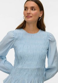 Šviesiai mėlyna palaidinė su reljefiniu, suraukytu audiniu, apvalia iškirpte ir pūstomis permatomomis rankovėmis. Turi laisvo kritimo siluetą ir subtilų blizgesį.