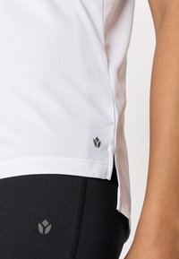 Weißes, strukturiertes Sporttop mit Schlitzen an den Seiten, das ein kleines graues Logo nahe dem Saum zeigt. Kombiniert mit schwarzen Leggings, die ein ähnliches Logo tragen.