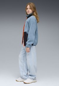 Fleecejacka i ljusblått med orange dragkedja och marinblå detaljer, ihop med lösa ljusblå jeans och vita sneakers. Slät textur.