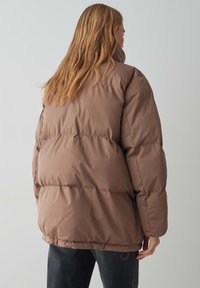 Giacca invernale marrone imbottita con colletto alto, caratterizzata da un design trapuntato, silhouette voluminosa ed orlo elastico.