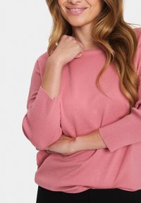Rosa gerippter Strickpullover mit dreiviertellangen Ärmeln, runder Halslinie und lockerer Passform, lässig über einem schwarzen Unterteil getragen.