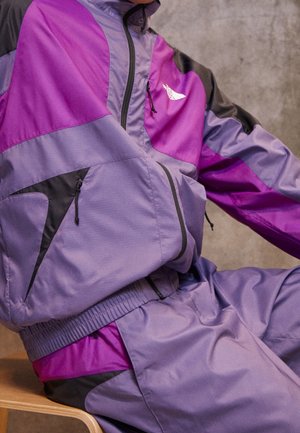 Personne assise portant une veste coupe-vent violet et violet avec un pantalon de survêtement avec des détails de fermeture éclair noirs et une ceinture élastique.