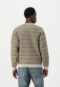 Sweater i olivengrøn med et struktureret, vævet mønster. Har en ribbet krave og manchetter og er parret med lyseblå jeans.