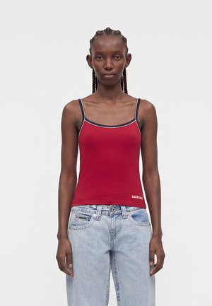 Woman with braided hair wearing a red spaghetti strap top with navy trim and light blue high-waisted jeans, standing against a plain background.