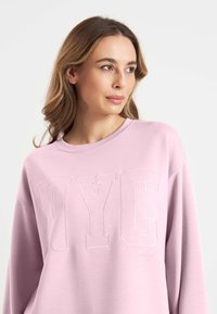 Lichtroze sweatshirt met "NYC" geborduurd in het wit, ronde halslijn en lange mouwen. Zachte textuur met een ontspannen pasvorm en minimale detaillering.