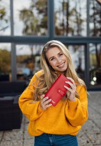 Red leather wallet with a snap closure and logo detail. Model in a bright yellow sweater holds it, set against a glass-windowed background.