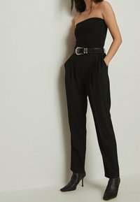 Black strapless top paired with high-waisted black trousers. Trousers feature pleats and pockets, complemented by a wide black belt and heeled boots.