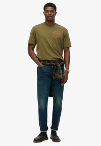 Camiseta de algodón verde oliva con logo rojo, jeans de denim azul y cinturón de cuero marrón, con una camisa de cuadros atada a la cintura. Zapatos negros.