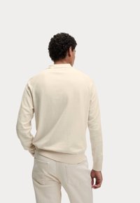 Mann mit lockigem Haar, der ein off-white Langarmhemd und passende Hose trägt, steht mit dem Rücken zu einem einfarbigen Hintergrund.