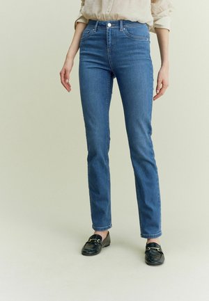 Femme portant un jean slim bleu, une chemise beige à manches longues retroussées et des mocassins noirs, debout sur un fond uni clair.