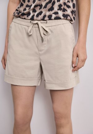 Beigenväriset nyörillä kiristettävät shortsit, joissa on käännetyt helmaukset, ja henkilö pitää päällään osittain shorteista näkyvää leopardi-kuvioista yläosaa.