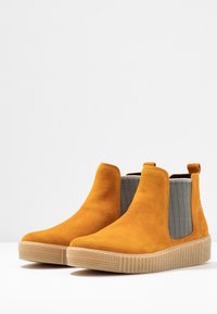 Bottes chevilles en daim jaune avec panneaux latéraux élastiques, tissu gris texturé et semelle en caoutchouc rainurée. Conçues avec une languette pour un enfilage facile.