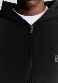 Felpa nera con zip, caratterizzata da una texture liscia, con zip frontale e un piccolo emblema sul lato sinistro del petto. Tessuto leggero, design casual.