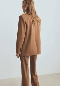 Traje marrón a medida con un corte holgado, que presenta un abrigo largo y pantalones de pernera recta. El material parece suave; incluye detalles con botones.