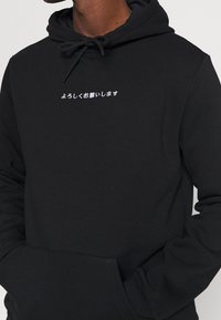 Svart hoodie i mjukt material, med en framficka och en textgrafik i vita japanska tecken över bröstet.