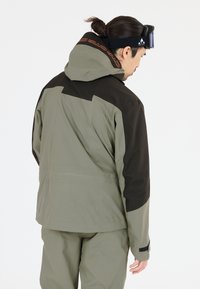 En vandtæt skijakke i olivengrøn og sort. Har en hætte, justerbare manchetter og et slankt, moderne design med en tekstureret overflade.