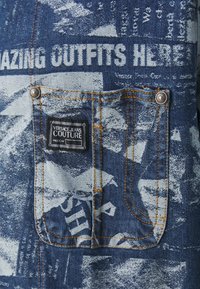 Poche en denim avec surpiqûres orange sur tissu bleu à motifs, avec une étiquette noire Versace Jeans Couture et un texte blanc indiquant "AMAZING OUTFITS HERE."