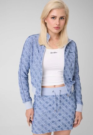 Femme blonde portant une veste zippée à motifs bleu clair et une jupe assortie avec un débardeur blanc court sur un fond uni.