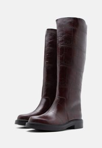 Burgundijas krūšu augstuma zābaki ar teksturētu krokodila ādas virsu un plakanām gumijas zolēm, ar gludu noapaļotu pirkstu dizainu.