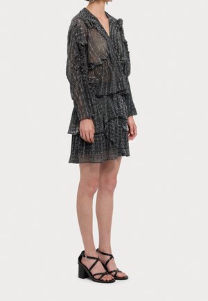 Femme vêtue d'une robe noire transparente à manches longues avec des volants, une jupe à étages et des sandales noires à talon bloc avec brides, debout sur un fond blanc.
