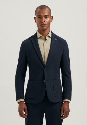 Marineblauwe blazer met een slim fit, twee voorkruiszakken en een subtiele textuur. Gecombineerd met een lichtbeige shirt met knoopdetails.