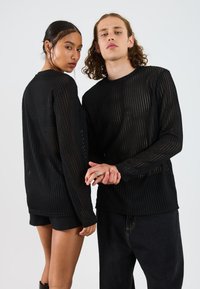 Two young adults wearing black mesh long-sleeve tops hold hands and look at the camera against a plain white background.