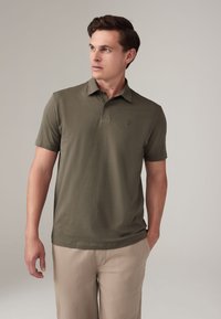 Polo verde oliva realizzato in tessuto morbido e traspirante; presenta un colletto classico, una chiusura con tre bottoni e un logo discreto sul petto.
