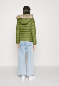 Oliven grøn puffjakke med en faux fur hætte, quiltet design og ribkantede manchetter. Sammen med vide lyseblå jeans og hvide sneakers.