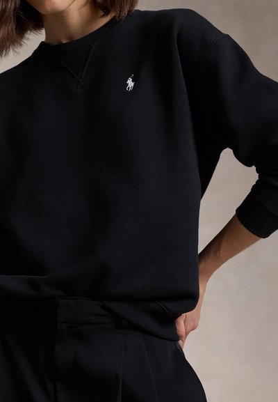 Inimene kannab musta pikakäistelist särk, millel on rindkeres väike valge Polo Ralph Laureni logo, ja musti pükse.